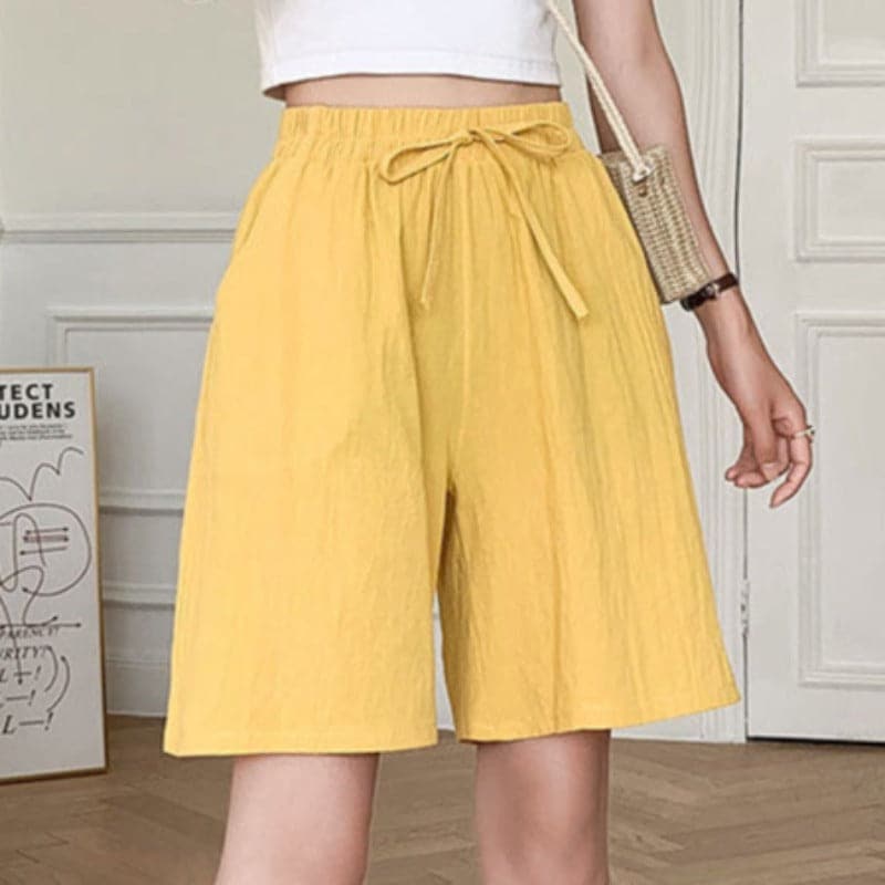 Short jaune femme avec ceinture nouée, style décontracté, devant une porte blanche. Mode estivale, tendance, confortable.