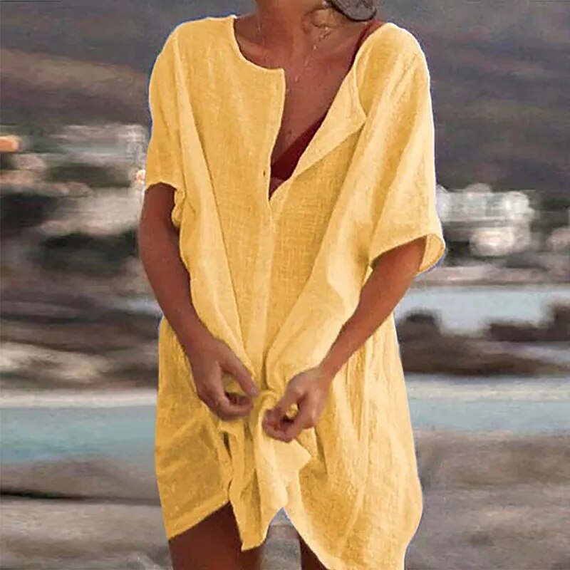 Robe de plage jaune en lin, manches courtes, portée par une femme sur fond de paysage côtier flou. Mode estivale, tenue décontractée.