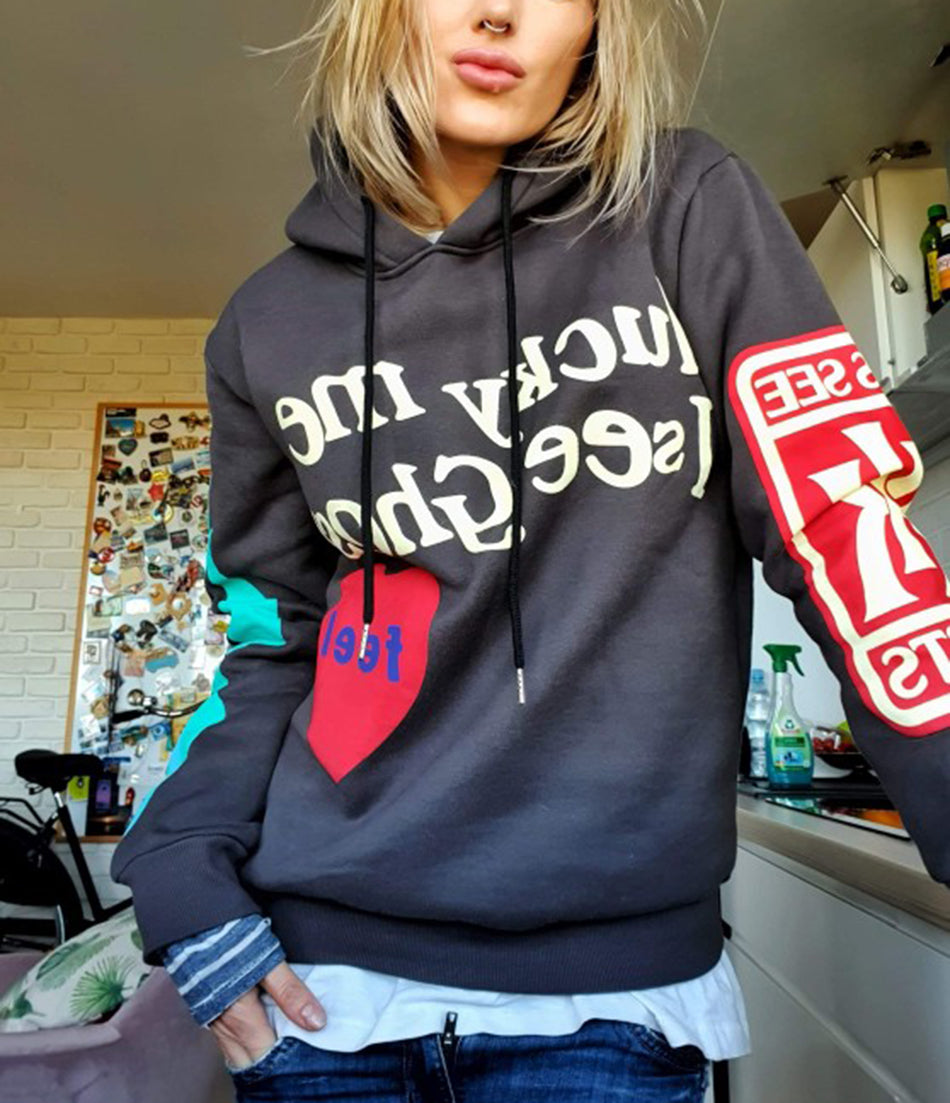 Femme portant un sweat à capuche gris avec motifs colorés, cœur rouge et texte. Style décontracté, mode urbaine, intérieur moderne.