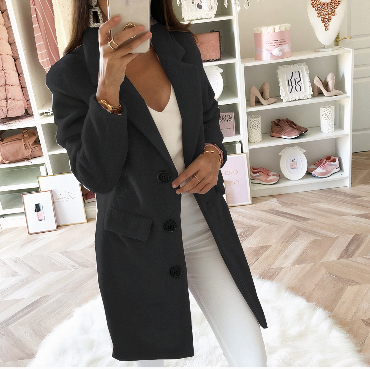 Femme en manteau noir et pantalon blanc prenant un selfie dans un dressing moderne avec étagères de sacs et chaussures, ambiance chic et élégante.