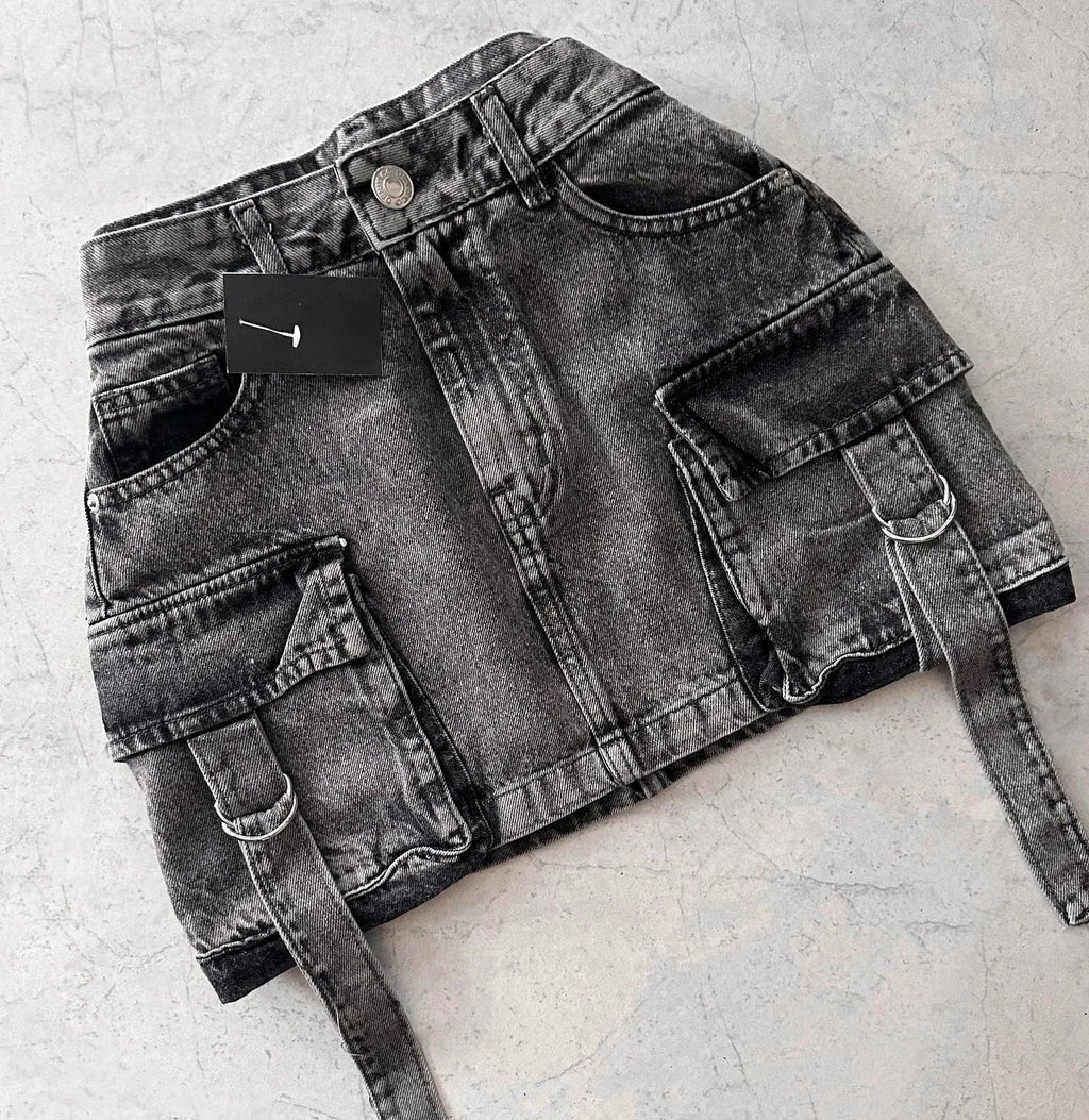 Mini-jupe en jean noir délavé avec poches cargo et ceinture, posée sur fond gris. Mode urbaine, tendance streetwear, style décontracté.