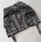Mini-jupe en jean noir délavé avec poches cargo et ceinture, posée sur fond gris. Mode urbaine, tendance streetwear, style décontracté.
