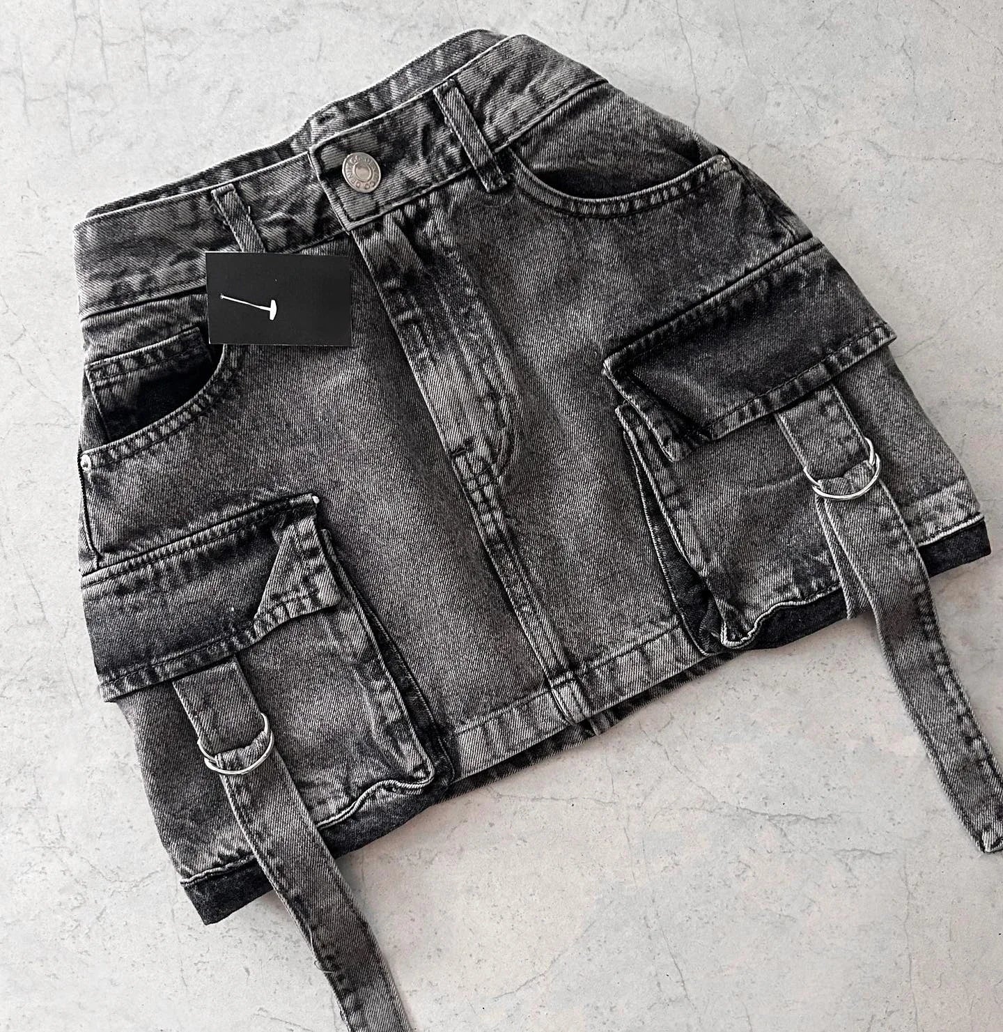 Mini-jupe en jean noir délavé avec poches cargo et ceinture, posée sur fond gris. Mode urbaine, tendance streetwear, style décontracté.
