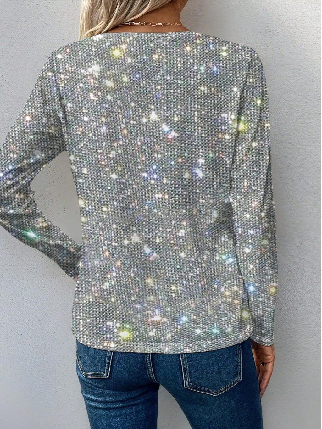 Femme portant un pull scintillant à manches longues, motif paillettes multicolores, dos tourné, avec un jean bleu. Mode tendance, style glamour.