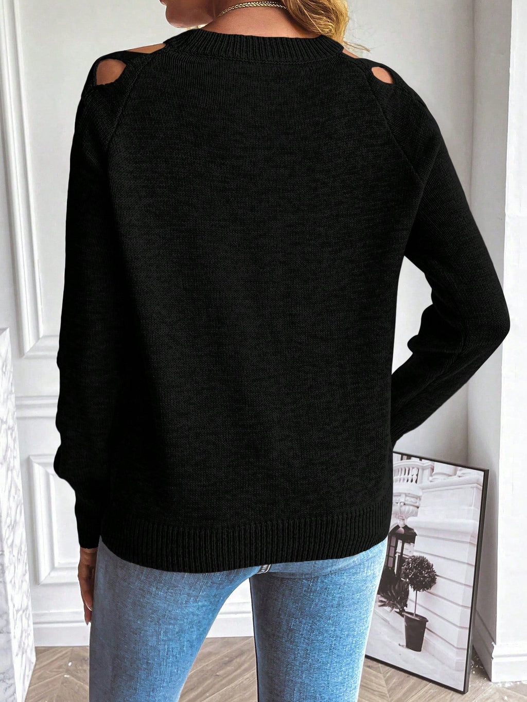 Femme portant un pull noir à découpes sur les épaules, associé à un jean bleu, dans un intérieur moderne. Mode automne-hiver, style décontracté.