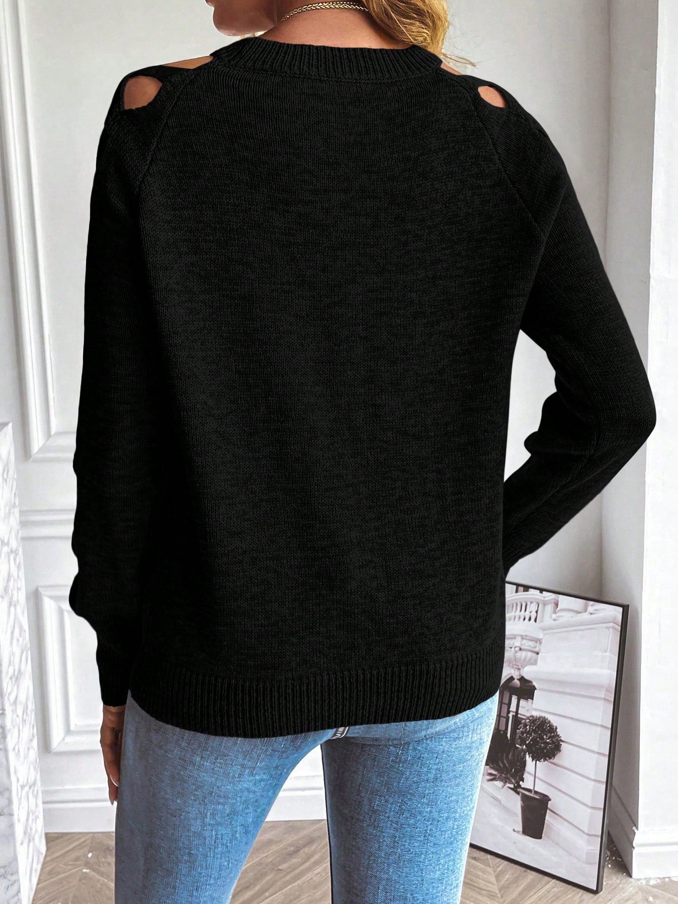 Femme portant un pull noir à découpes sur les épaules, associé à un jean bleu, dans un intérieur moderne. Mode automne-hiver, style décontracté.