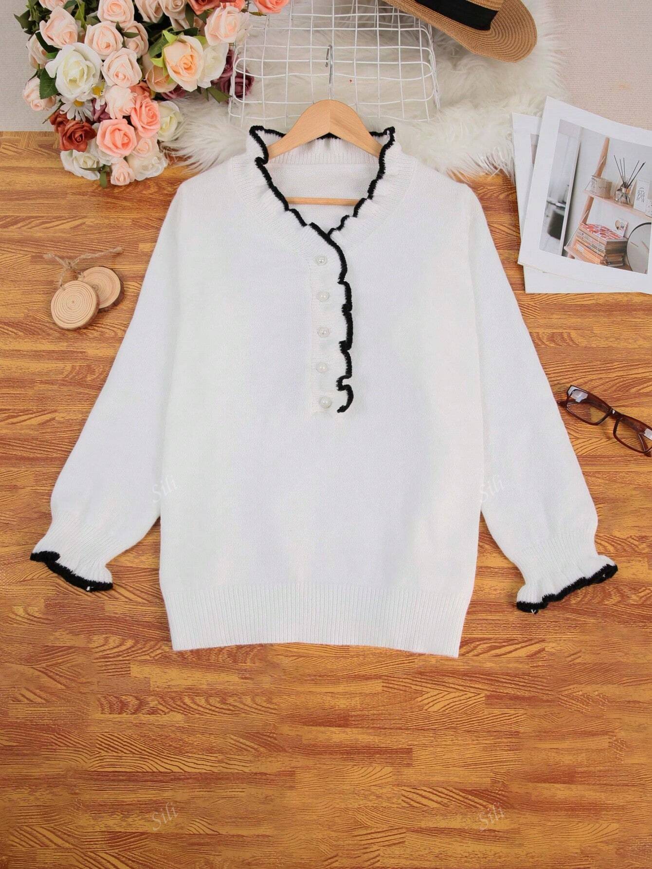 Pull blanc à col volanté noir, posé sur une table en bois avec des lunettes, des fleurs et des magazines. Mode féminine élégante et tendance.