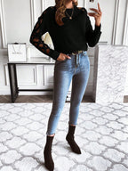 Femme en pull noir à manches ajourées, jeans slim bleu, bottines noires, posant dans un intérieur moderne avec tapis géométrique et décor minimaliste.