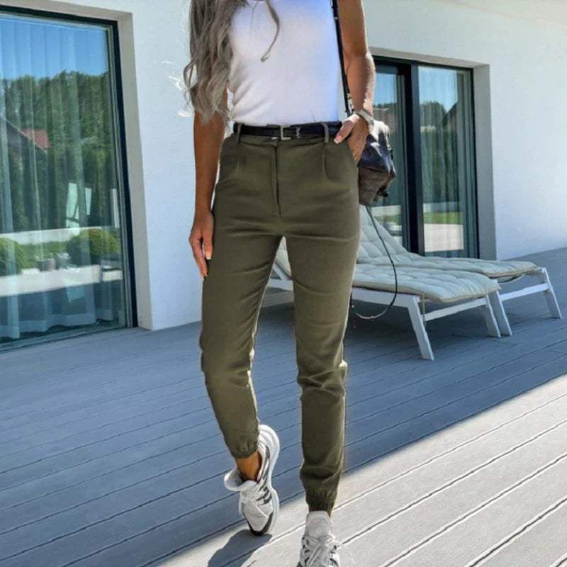 Femme en pantalon kaki taille haute, t-shirt blanc, baskets blanches, posant sur une terrasse moderne avec chaise longue et grandes fenêtres.