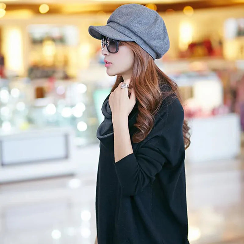 Femme élégante portant un chapeau gris et des lunettes de soleil, vêtue d'un pull noir, dans un centre commercial lumineux. Mode automne chic.