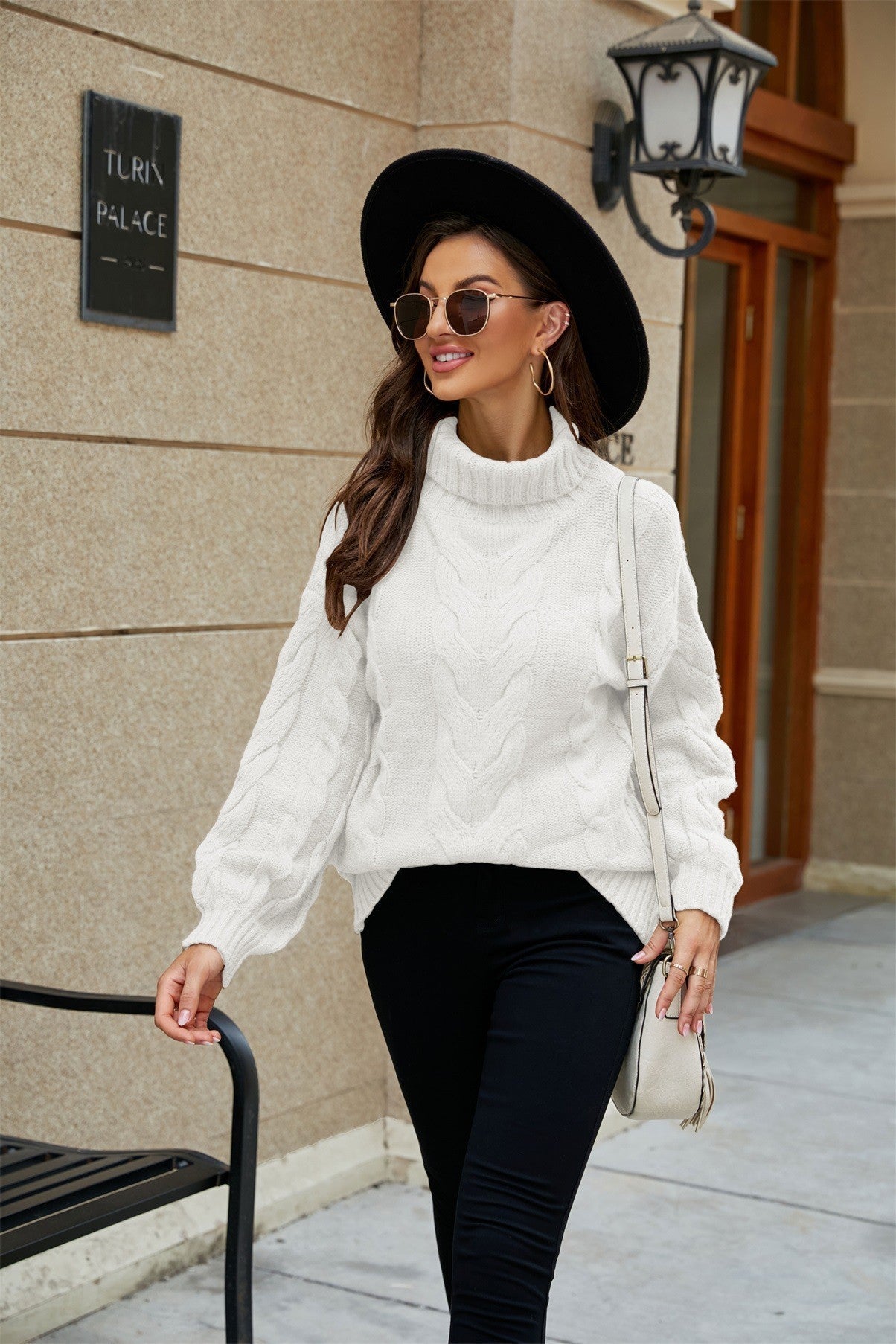 Femme élégante portant un pull blanc torsadé, chapeau noir et lunettes de soleil, marchant en ville. Mode automne, style urbain chic.