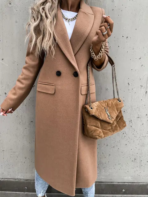 Femme portant un manteau camel élégant, sac à main en daim marron, chaîne dorée, sur fond de mur gris. Mode automne chic, tendance urbaine.