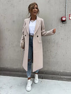 Femme en manteau beige élégant, jeans déchirés et baskets blanches, posant contre un mur en béton. Mode automne chic, style urbain décontracté.