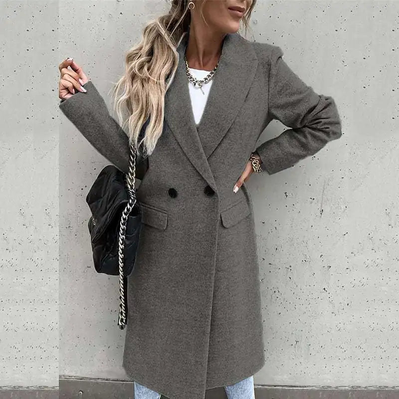 Femme portant un manteau gris élégant, sac noir à chaîne, cheveux ondulés, mur en béton en arrière-plan. Mode automne chic, tendance urbaine.