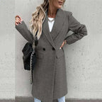 Femme portant un manteau gris élégant, sac noir à chaîne, cheveux ondulés, mur en béton en arrière-plan. Mode automne chic, tendance urbaine.
