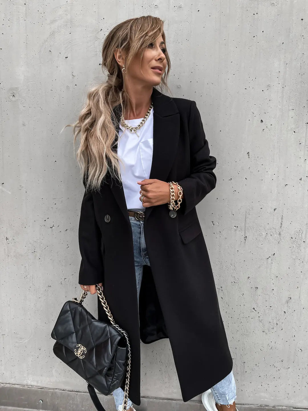 Femme élégante portant un manteau noir, t-shirt blanc, jeans déchirés, sac à main noir en cuir, posant contre un mur gris. Mode automne chic.