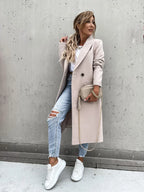 Femme en manteau beige, jean déchiré, baskets blanches, posant contre un mur gris avec un sac à main beige. Mode urbaine chic et décontractée.