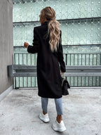 Femme de dos avec manteau noir, jeans, baskets blanches, sac à main noir, cheveux longs ondulés, devant une structure métallique moderne.