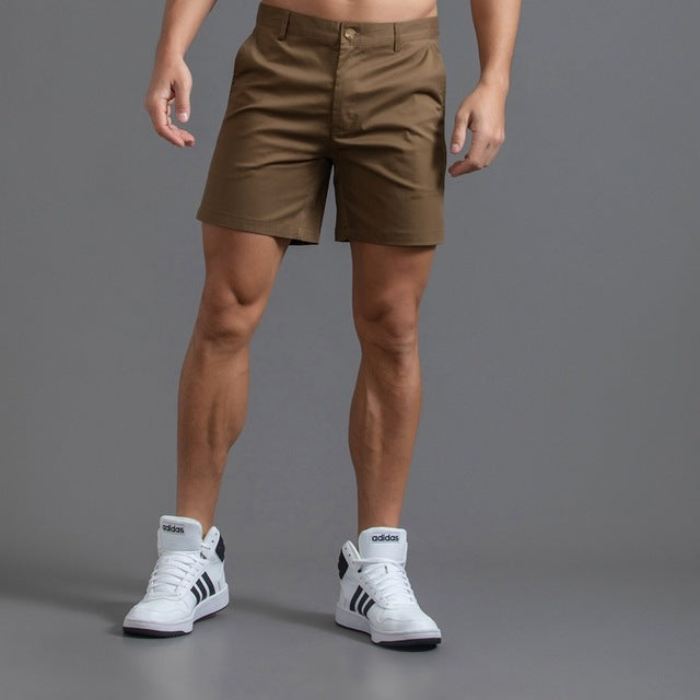 Homme portant un short marron et des baskets blanches Adidas sur fond gris. Mode masculine, tenue décontractée, style sportif.