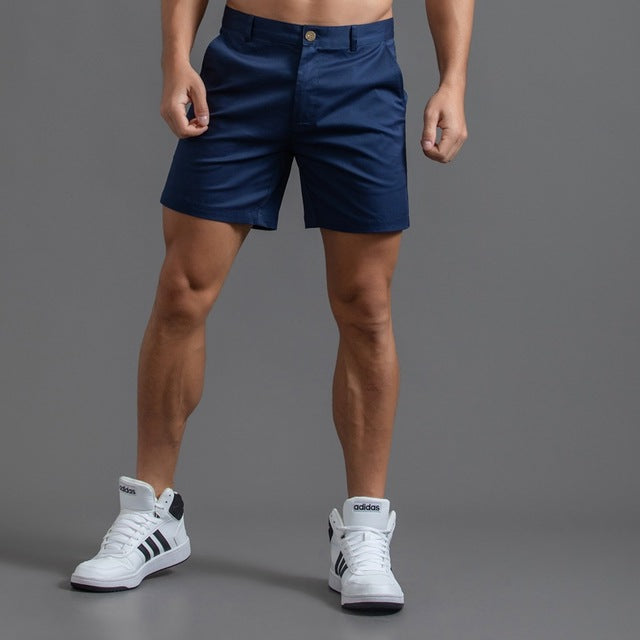 Homme portant un short bleu marine et des baskets blanches Adidas sur fond gris. Mode masculine, tenue décontractée, style sportif.