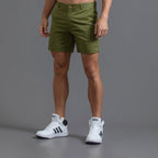Homme portant un short vert olive et des baskets blanches Adidas sur fond gris. Mode masculine, tenue décontractée, style sportif.