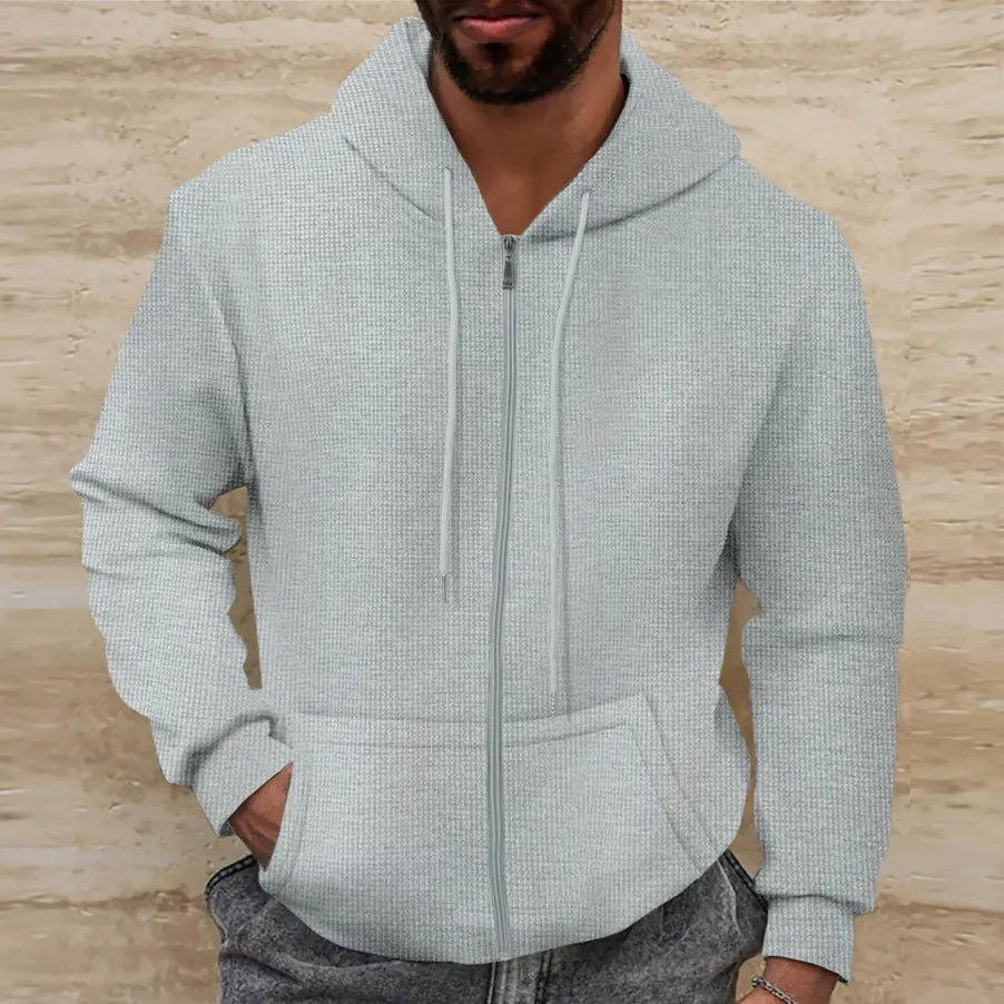 Homme portant un sweat à capuche gris clair avec fermeture éclair, mains dans les poches, devant un mur beige. Mode décontractée, vêtements pour hommes.