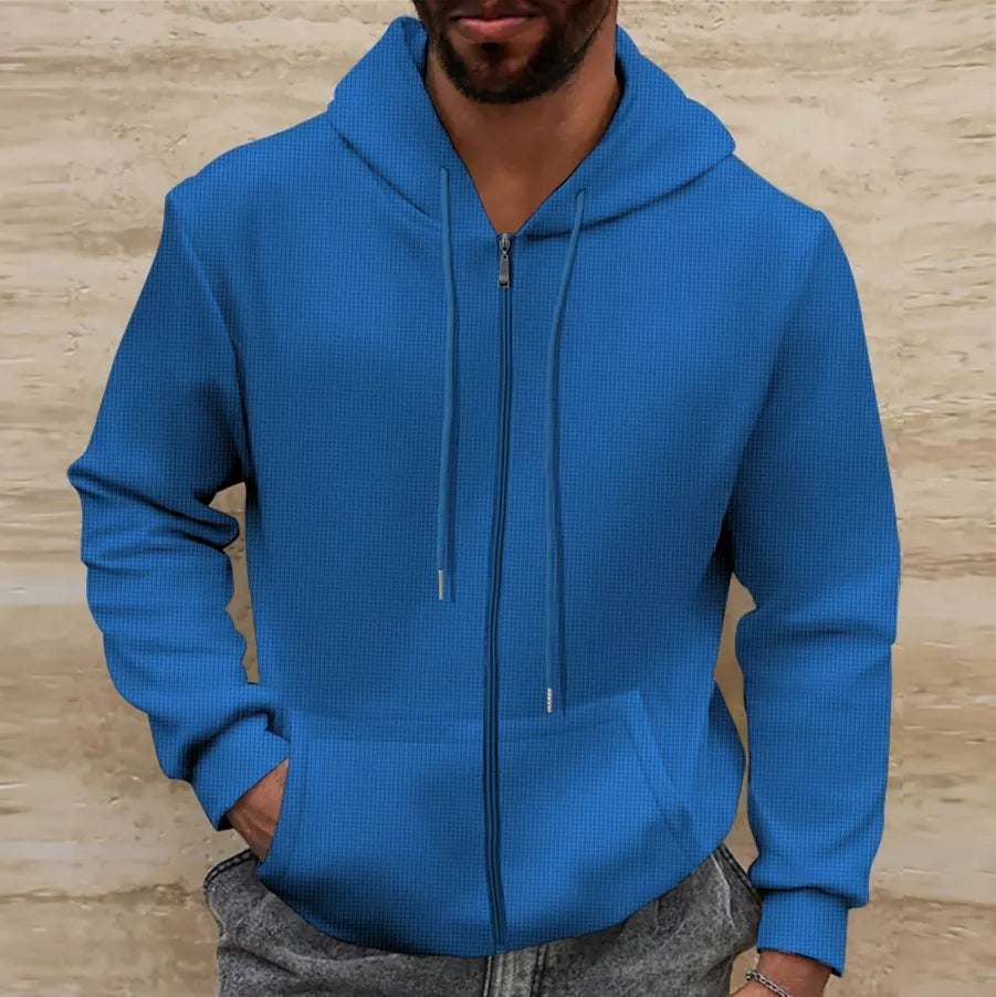 Homme portant un sweat à capuche bleu avec fermeture éclair, mains dans les poches, sur fond de mur beige. Mode décontractée, vêtement tendance.
