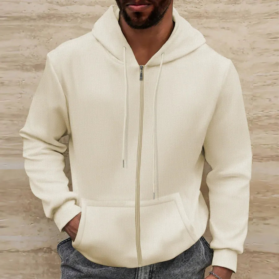 Homme portant un sweat à capuche beige, fermeture éclair, poches avant, sur fond de mur texturé. Mode décontractée, tendance automne-hiver.