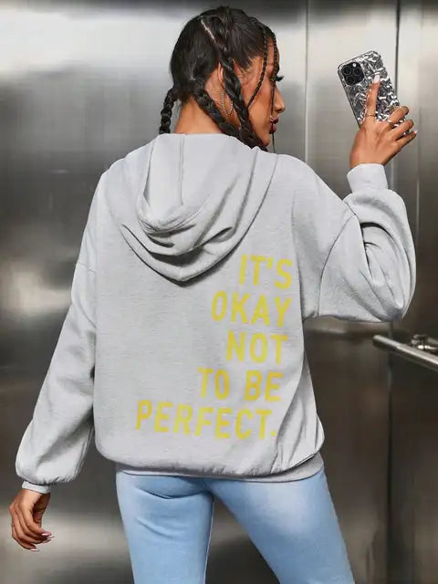 Femme de dos portant un sweat à capuche gris avec le texte "It's okay not to be perfect", tenant un smartphone, dans un ascenseur moderne.