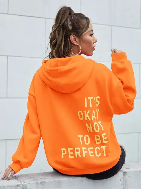Femme portant un sweat à capuche orange avec le texte "It's okay not to be perfect", posant de dos. Mode décontractée, tendance automne.