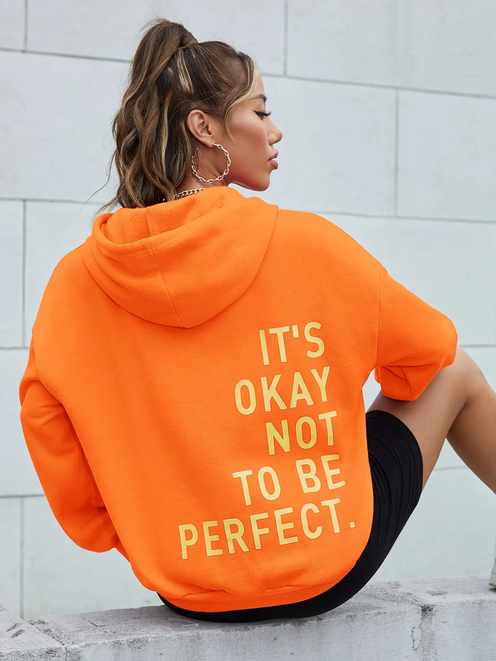 Femme portant un sweat à capuche orange avec le texte "It's okay not to be perfect", assise contre un mur blanc. Mode décontractée, style urbain.