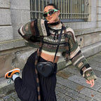 Femme posant en pull tricoté multicolore, jupe noire, lunettes de soleil vertes et sac en forme de cœur, devant un bâtiment en pierre.