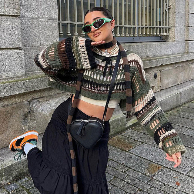 Femme posant en pull tricoté multicolore, jupe noire, lunettes de soleil vertes et sac en forme de cœur, devant un bâtiment en pierre.