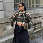 Femme portant un pull en maille multicolore, jupe noire longue, lunettes de soleil vertes, sac noir, posant devant un mur en pierre. Mode urbaine chic.