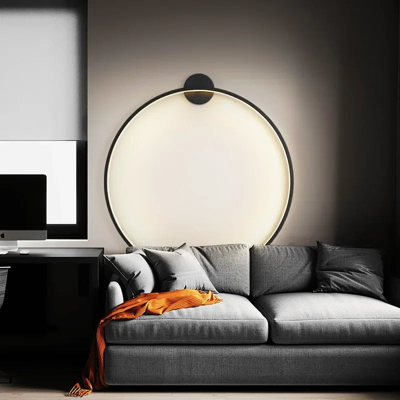 Canapé gris moderne avec coussin orange, éclairé par une lampe murale circulaire LED, dans un salon minimaliste avec bureau noir et fenêtre.