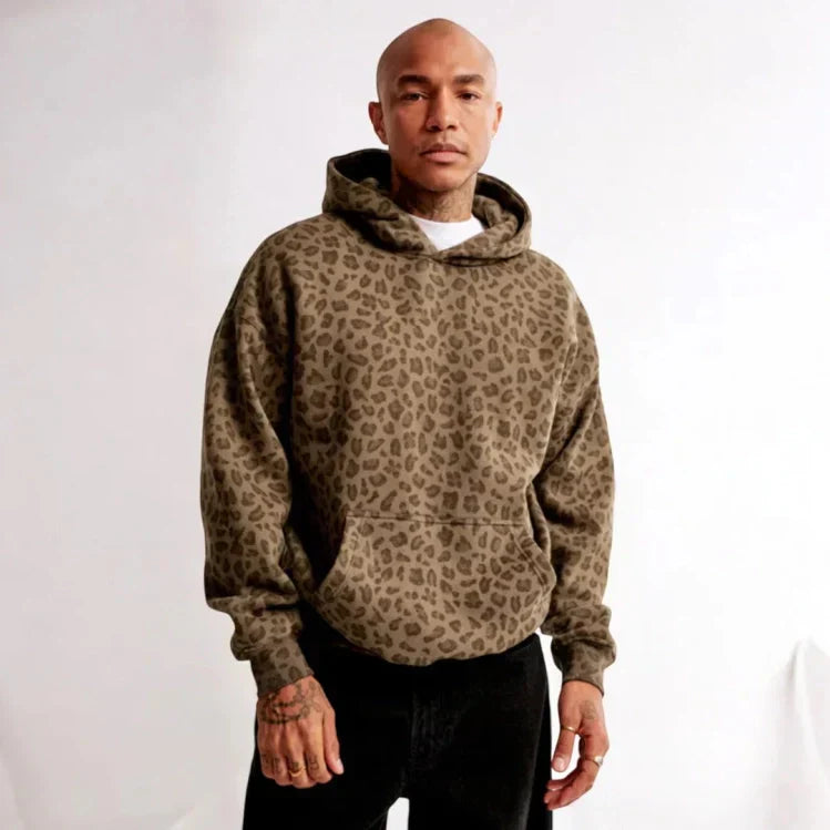Homme portant un sweat à capuche imprimé léopard marron, mode urbaine tendance, style décontracté, fond blanc.