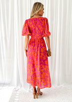 Robe longue fleurie rose et orange, manches bouffantes, portée par une femme blonde de dos, tenant une pochette marron, sur fond de rideaux blancs.