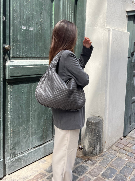 Femme de dos portant un sac à main tressé noir, veste grise, pantalon blanc, devant une porte verte en bois, rue pavée. Mode urbaine chic.