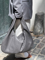 Sac à main tissé gris porté par une personne en manteau gris et pantalon blanc, sur un trottoir pavé. Mode urbaine, accessoire tendance.