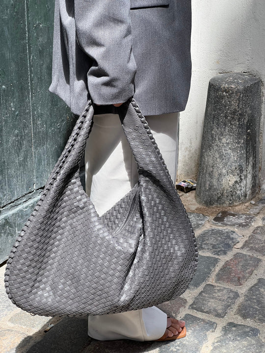 Sac à main tissé gris porté par une personne en manteau gris et pantalon blanc, sur un trottoir pavé. Mode urbaine, accessoire tendance.