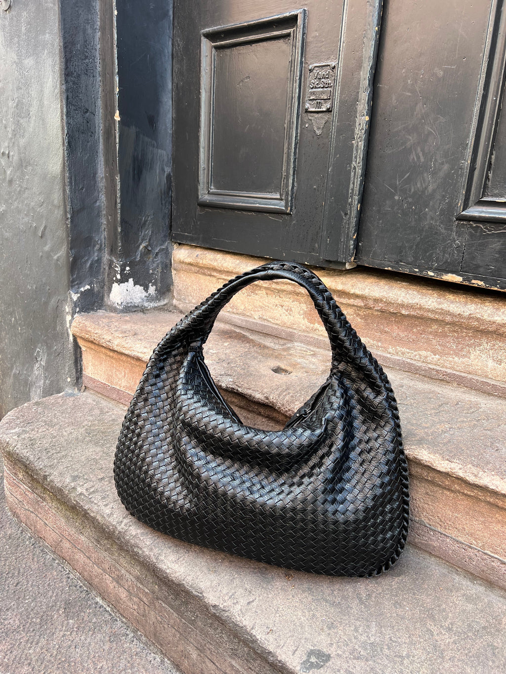 Sac à main en cuir tressé noir posé sur des marches en pierre devant une porte noire. Accessoire mode élégant et tendance.