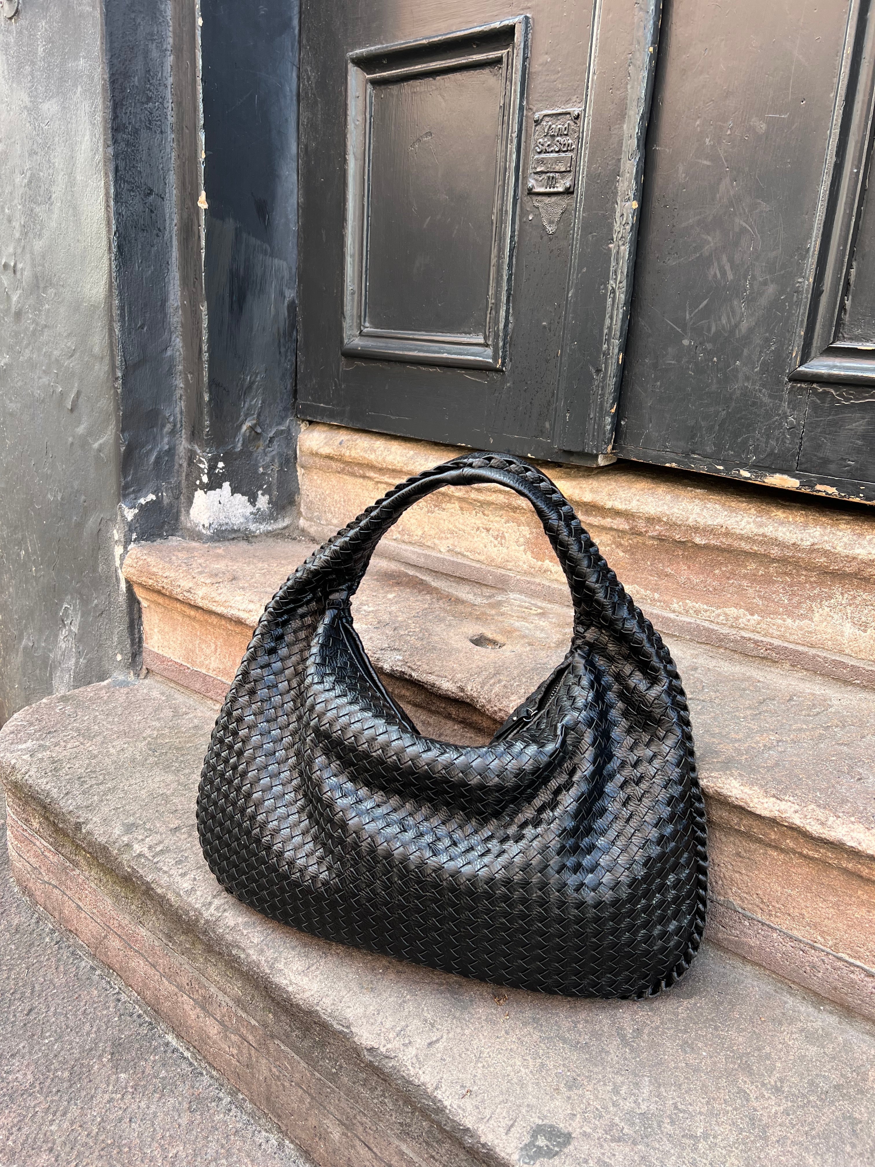 Sac à main en cuir tressé noir posé sur des marches en pierre devant une porte noire. Accessoire mode élégant et tendance.