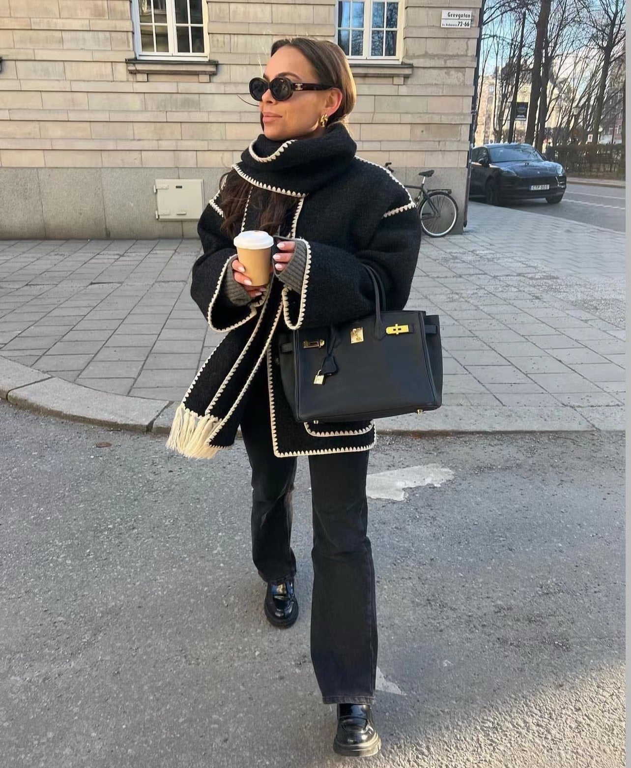 Femme élégante en manteau noir avec écharpe, lunettes de soleil, tenant un café et un sac à main de luxe, marchant en ville sur un trottoir pavé.