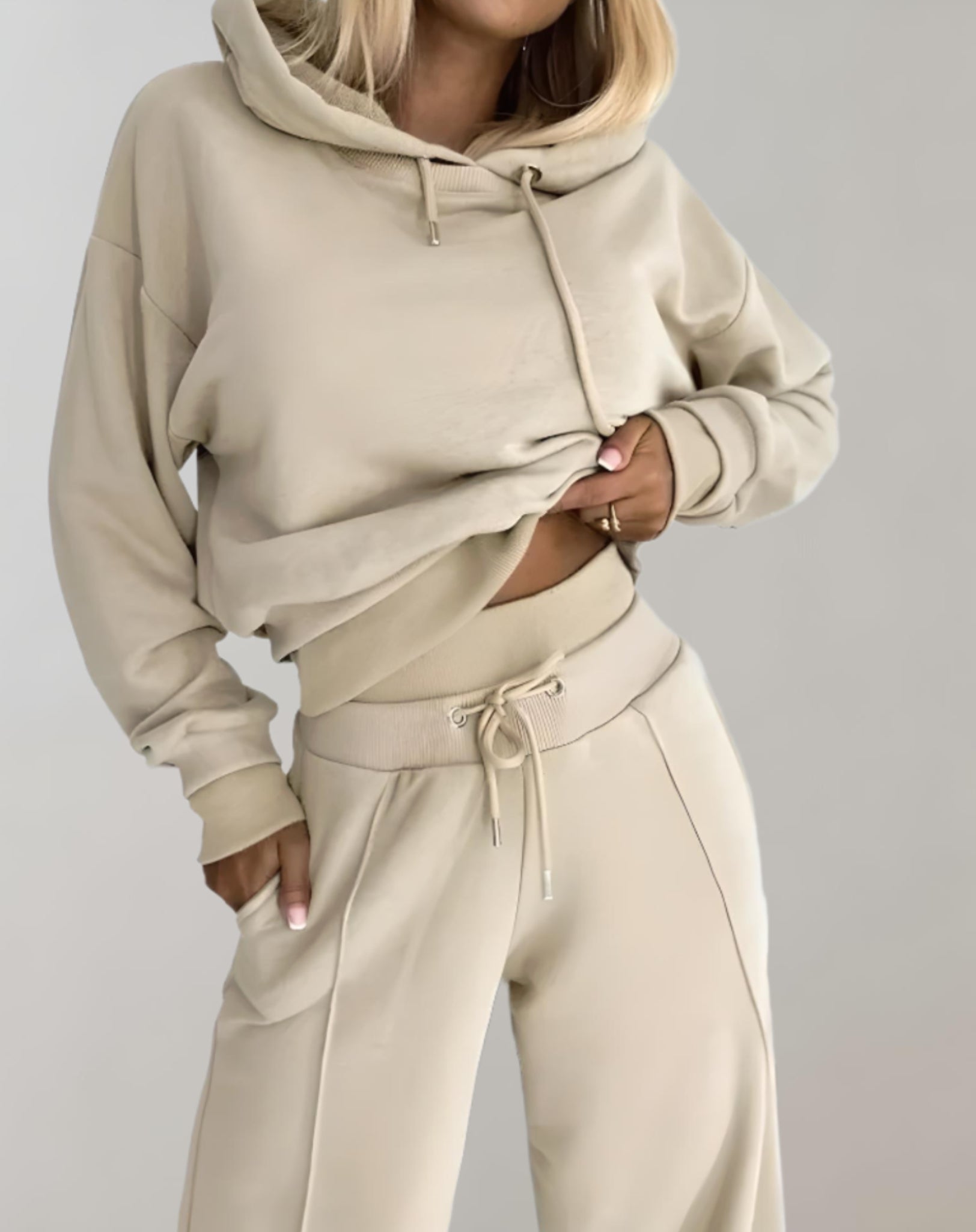 Femme portant un ensemble de survêtement beige, composé d'un sweat à capuche et d'un pantalon ample. Mode décontractée, tendance automne-hiver.