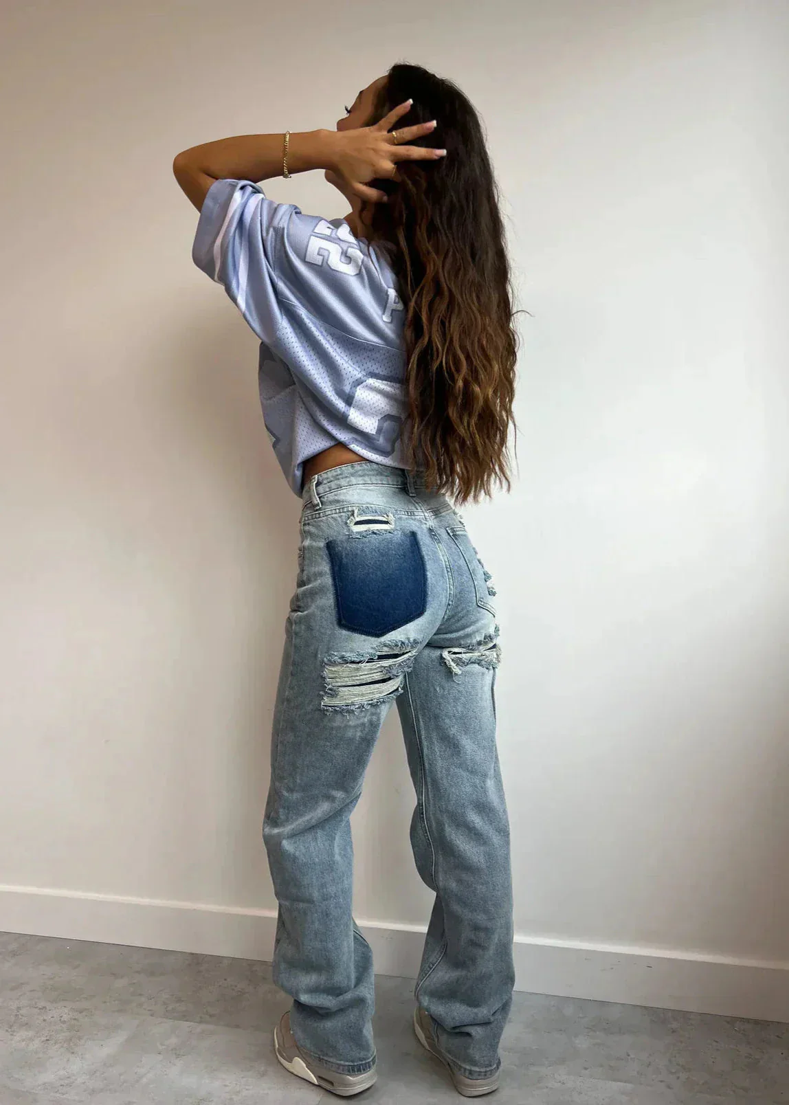 Femme de dos portant un jean déchiré et un t-shirt ample, posant contre un mur blanc. Mode décontractée, style urbain, tendance denim.