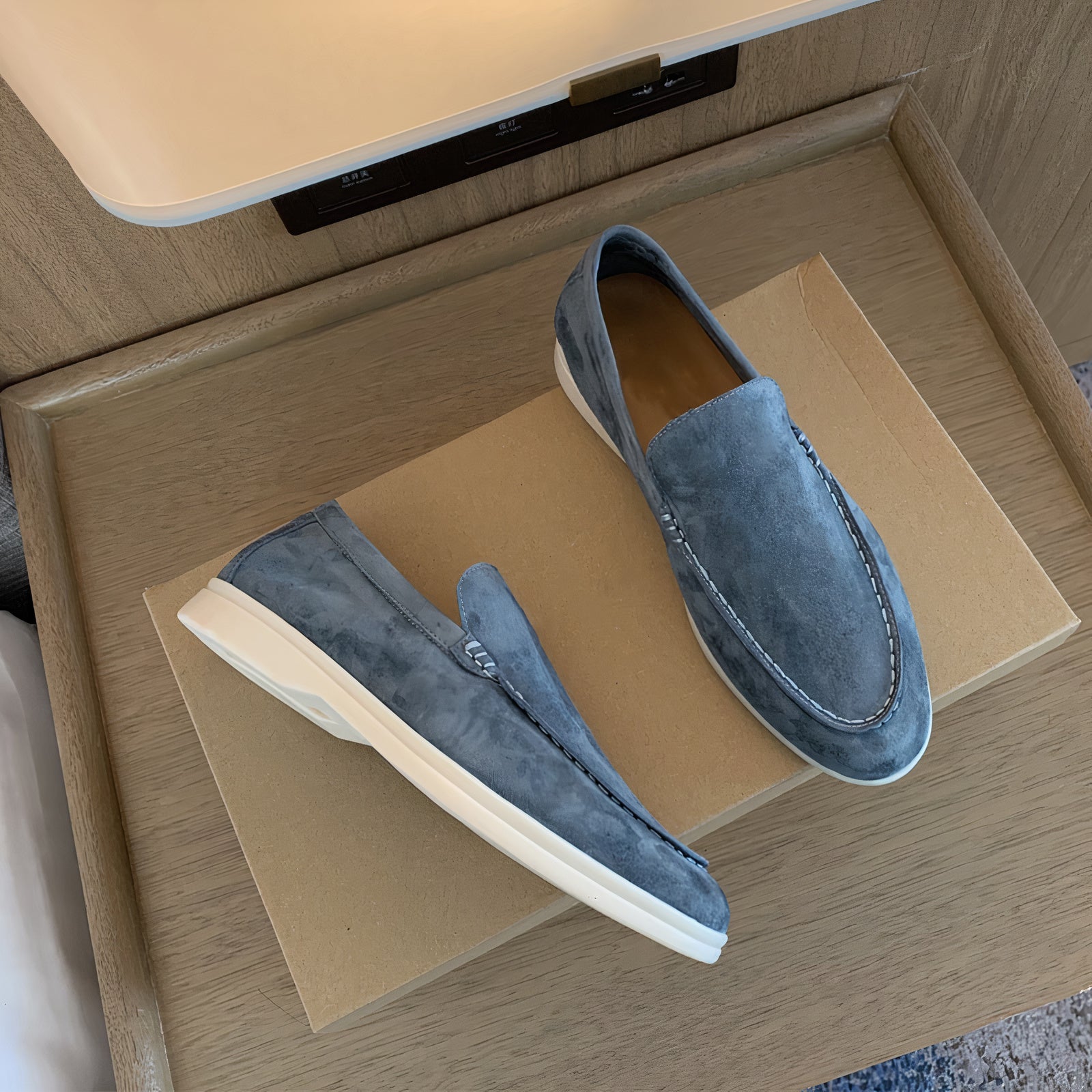 Mocassins en daim bleu sur boîte en carton, design élégant et moderne, semelle blanche contrastante, chaussures pour homme tendance et confortables.