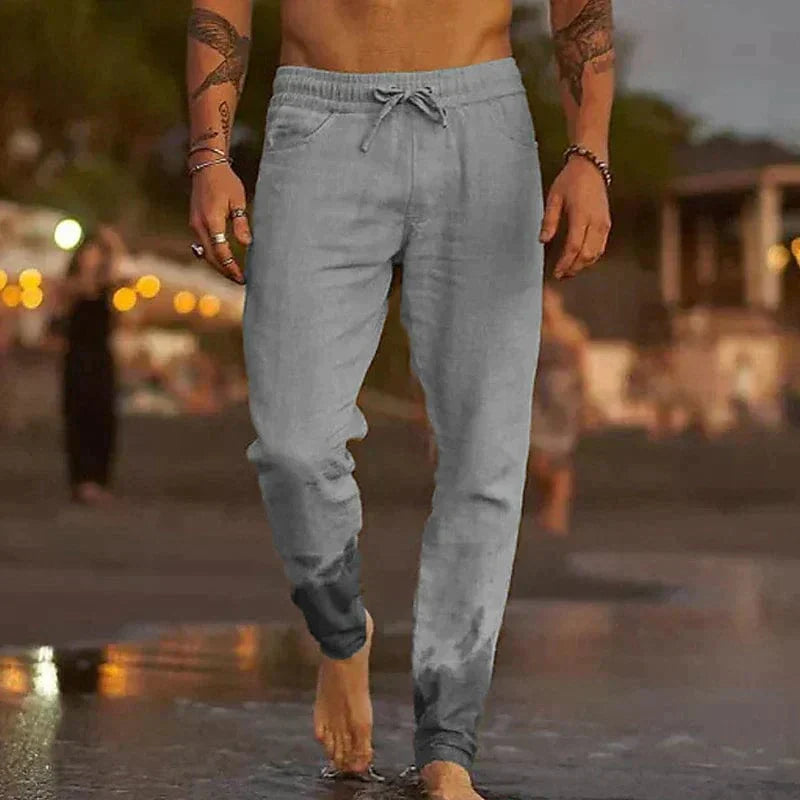 Homme marchant sur la plage au coucher du soleil, portant un pantalon de jogging gris, pieds nus dans l'eau, ambiance détendue et estivale.