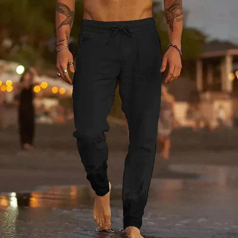 Homme marchant sur la plage au crépuscule, portant un pantalon de jogging noir. Scène estivale, ambiance décontractée, bord de mer.