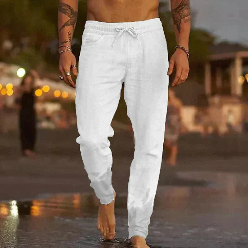 Homme marchant sur la plage au coucher du soleil, portant un pantalon de jogging blanc. Mode décontractée, ambiance estivale, détente bord de mer.