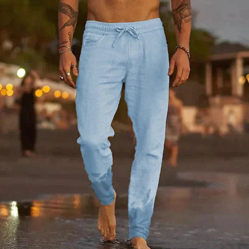 Homme marchant sur la plage au coucher du soleil, portant un pantalon de jogging bleu clair. Mode décontractée, style estival, ambiance bord de mer.